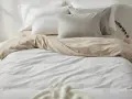 Ein Bett mit weißer und sandfarbener Bettwäsche aus Satin-Baumwolle. Mehrere Kissen liegen auf dem Bett.