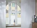 Weiße Schiebegardinen in alten Schienen vor einem Bogenfenster mit Blick auf ein Gebäude. Eine Puppe steht auf der Fensterbank.