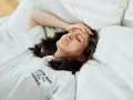 Frau liegt im Bett, Stirn gerunzelt, Hand an der Schläfe. Zu viele Gedanken im Kopf, kein Schlaf möglich.