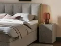 Gemütliches Queensize Bett mit grauem Stoffbezug, Kissen und Decke. Daneben ein Nachttisch mit Lampe.