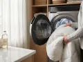 Eine Person legt eine weiße Daunendecke in eine Waschmaschine. Neben der Maschine steht eine Flasche mit Waschmittel.