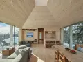 Gemütliches Wohnzimmer mit Holzwänden und Decke, die ein kariertes Muster andeuten. Große Fensterfronten bieten Blick auf verschneite Landschaft.