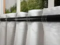 Weiße Polyestergardinen hängen glatt an einer dunklen Stange. Der Stoff fällt in weichen Falten.