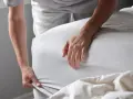 Eine Person zieht ein hellgraues **Bettlaken** über eine Matratze. Das Laken ist glatt und liegt gut an.