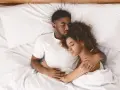 Ein junges Paar schläft eng zusammen in einem Bett. Er hält sie schützend im Arm, während sie sich an ihn kuschelt.
