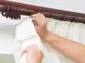 Hände befestigen weiße Gardinen an einer dunklen Holzstange mit Ringen.