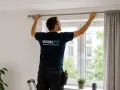 Mann montiert eine Gardinenstange über einem Fenster. Er steht auf einer Leiter und richtet die Stange aus, bevor er sie bohrt.