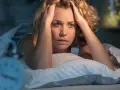 Verzweifelte Frau im Bett, Hände am Kopf, fragt sich, was die Wolfsstunde ist. Ein Wecker ist unscharf im Vordergrund zu sehen.