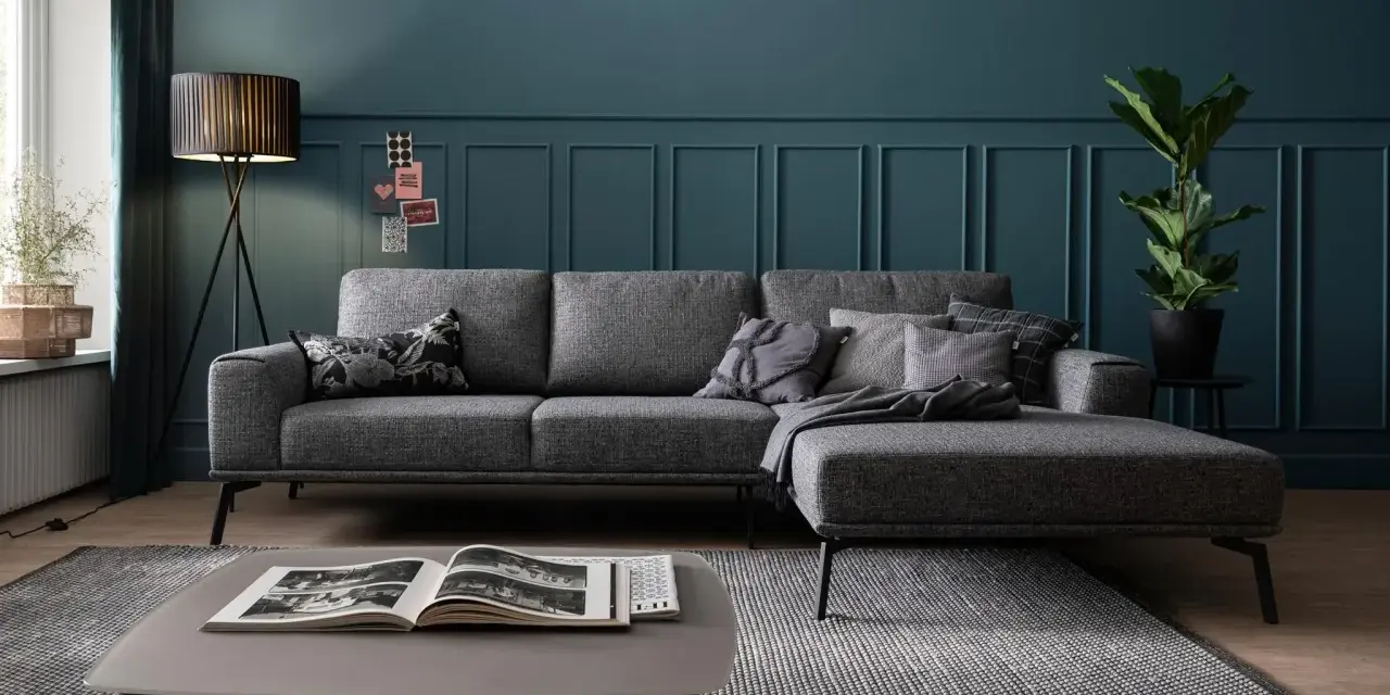 Ein moderner Wohnbereich mit einem anthrazitfarbenen Sofa, das auf einem gemusterten Teppich steht. Ein Couchtisch mit einem offenen Buch und eine Stehlampe vervollständigen das Bild.