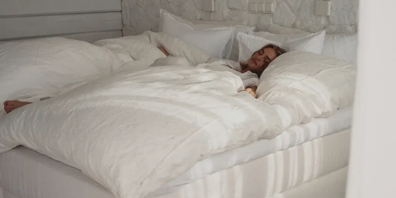 Eine Frau schläft tief unter einer frisch gewaschenen, weißen Bettdecke. Die Sonne wirft Streifen auf das Bett.