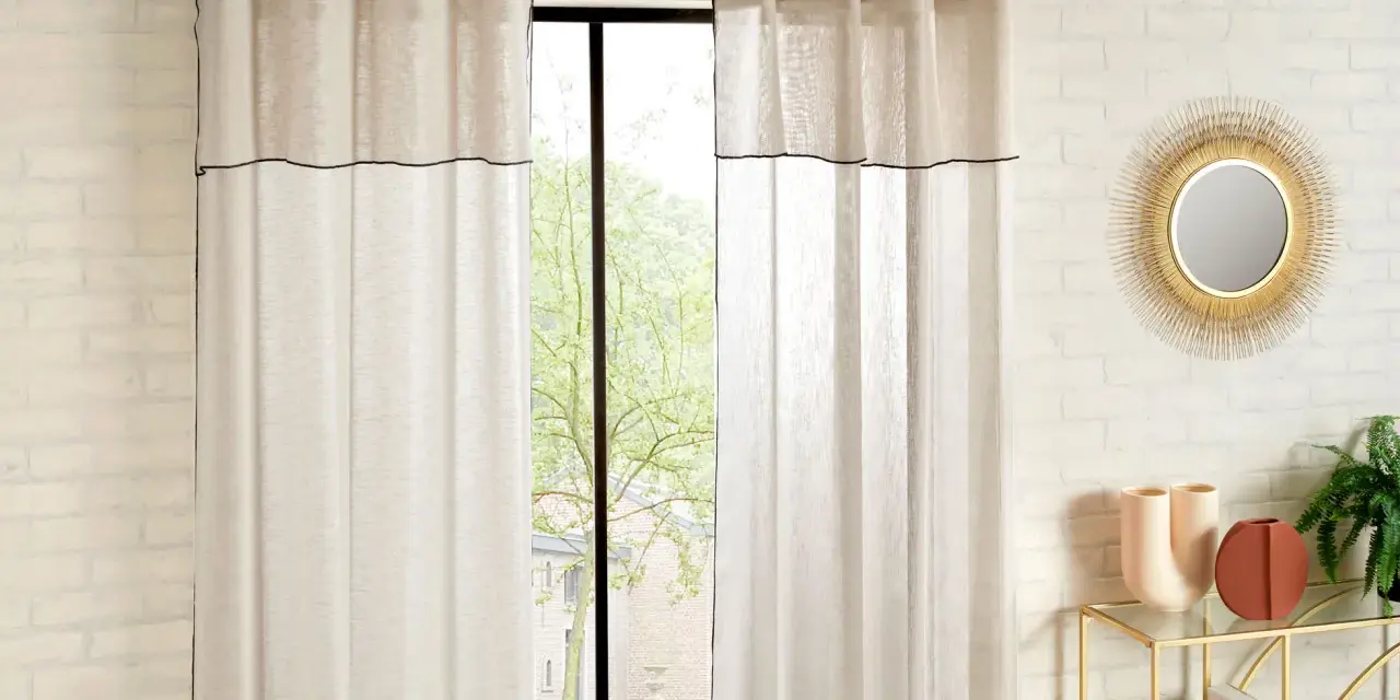 Helle Gardinen mit schwarzer Umrandung sind hochgesteckt und rahmen ein Fenster mit Blick ins Grüne ein.