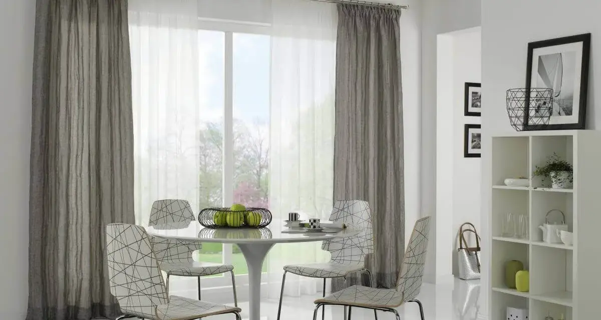 Ein Essbereich mit einem runden Tisch, vier Stühlen und einem Fenster, das von einer Gardinenschiene oder Stange mit grauen und weißen Vorhängen eingerahmt wird.