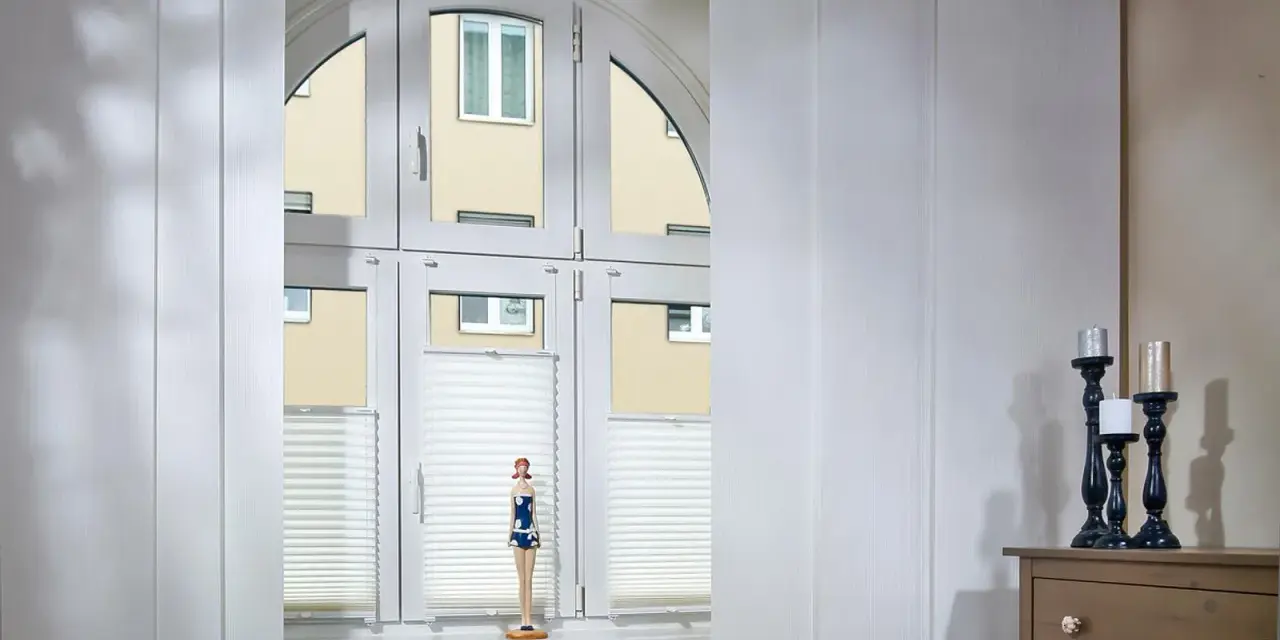 Weiße Schiebegardinen in alten Schienen vor einem Bogenfenster mit Blick auf ein Gebäude. Eine Puppe steht auf der Fensterbank.