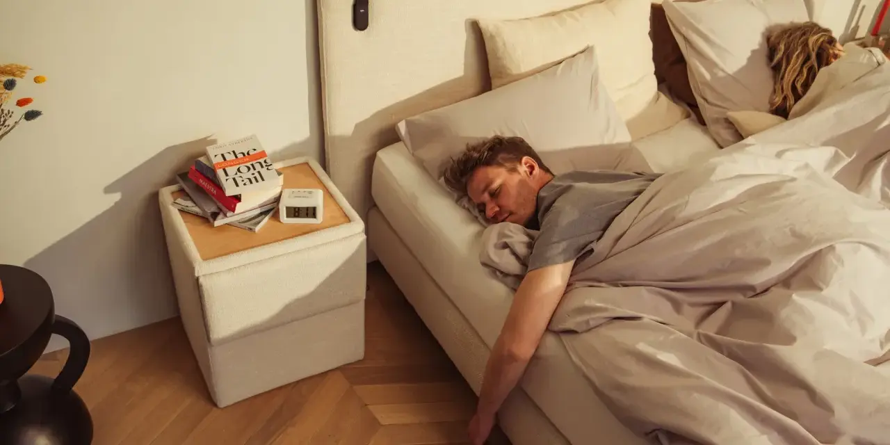 Mann schläft tief und fest auf einem Bett, das sich wie **Kaltschaum** anfühlt. Neben dem Bett liegen Bücher und ein Wecker.