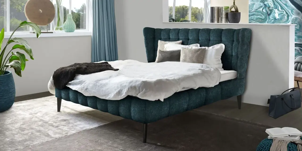 Ein luxuriöses Bett mit tiefblauem Polster und weißen Bettwäsche-Ideen. Dekorative Kissen und eine dunkle Decke laden zum Entspannen ein.