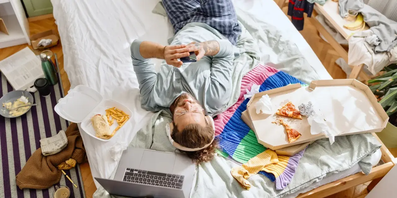 Mann liegt im Chaos auf dem Bett, umgeben von Essensresten und Kleidung. Ein typischer Tag des "bed rotting".