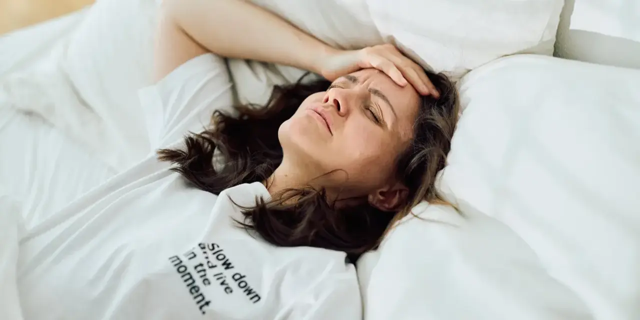 Frau liegt im Bett, Stirn gerunzelt, Hand an der Schläfe. Zu viele Gedanken im Kopf, kein Schlaf möglich.