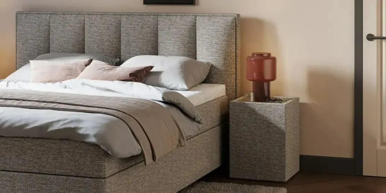 Gemütliches Queensize Bett mit grauem Stoffbezug, Kissen und Decke. Daneben ein Nachttisch mit Lampe.