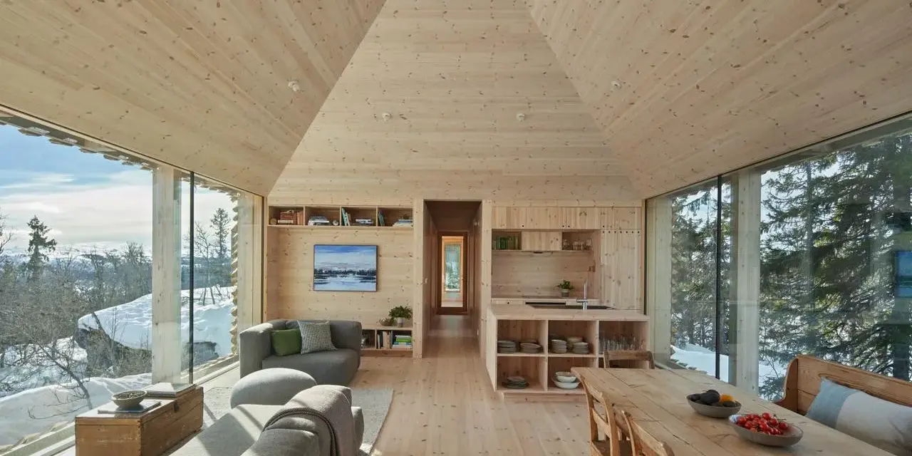 Gemütliches Wohnzimmer mit Holzwänden und Decke, die ein kariertes Muster andeuten. Große Fensterfronten bieten Blick auf verschneite Landschaft.