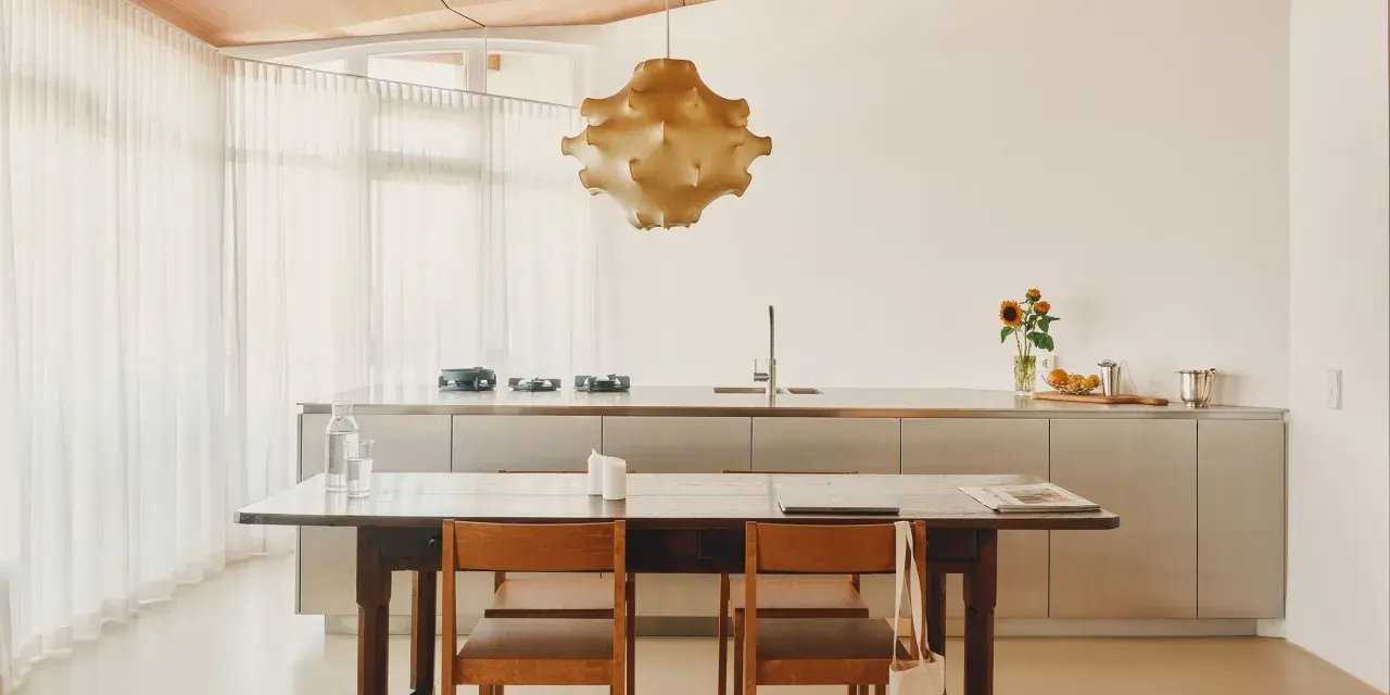 Ein Essbereich mit Holztisch und Stühlen, der den schwedischen Wohnstil verkörpert. Dahinter eine moderne Küche mit Edelstahlfronten und eine auffällige Hängelampe.
