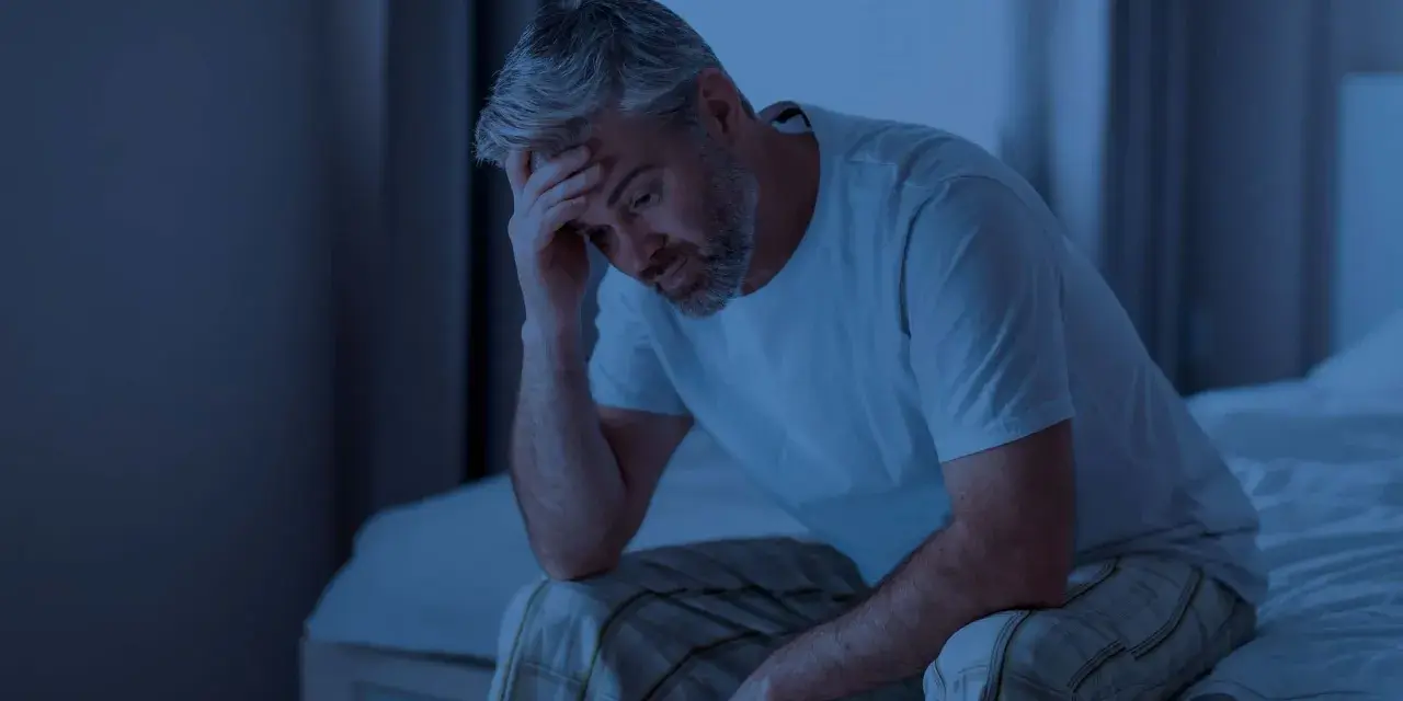 Ein Mann sitzt im Dunkeln auf seinem Bett, die Hand an der Stirn. Er fragt sich, wie lange man ohne Schlaf auskommt.