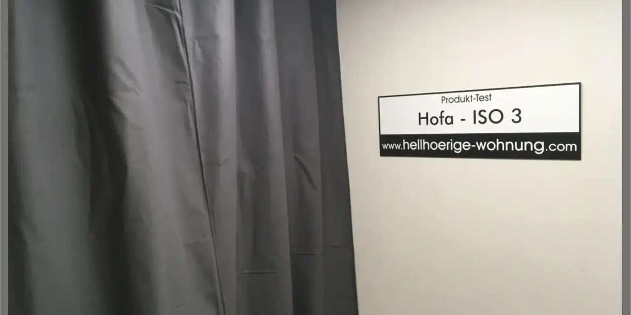 Grauer Schallschutzvorhang im Test. Ein Schild mit "Produkt-Test Hofa - ISO 3" und "www.hellhoerige-wohnung.com" hängt an der Wand.