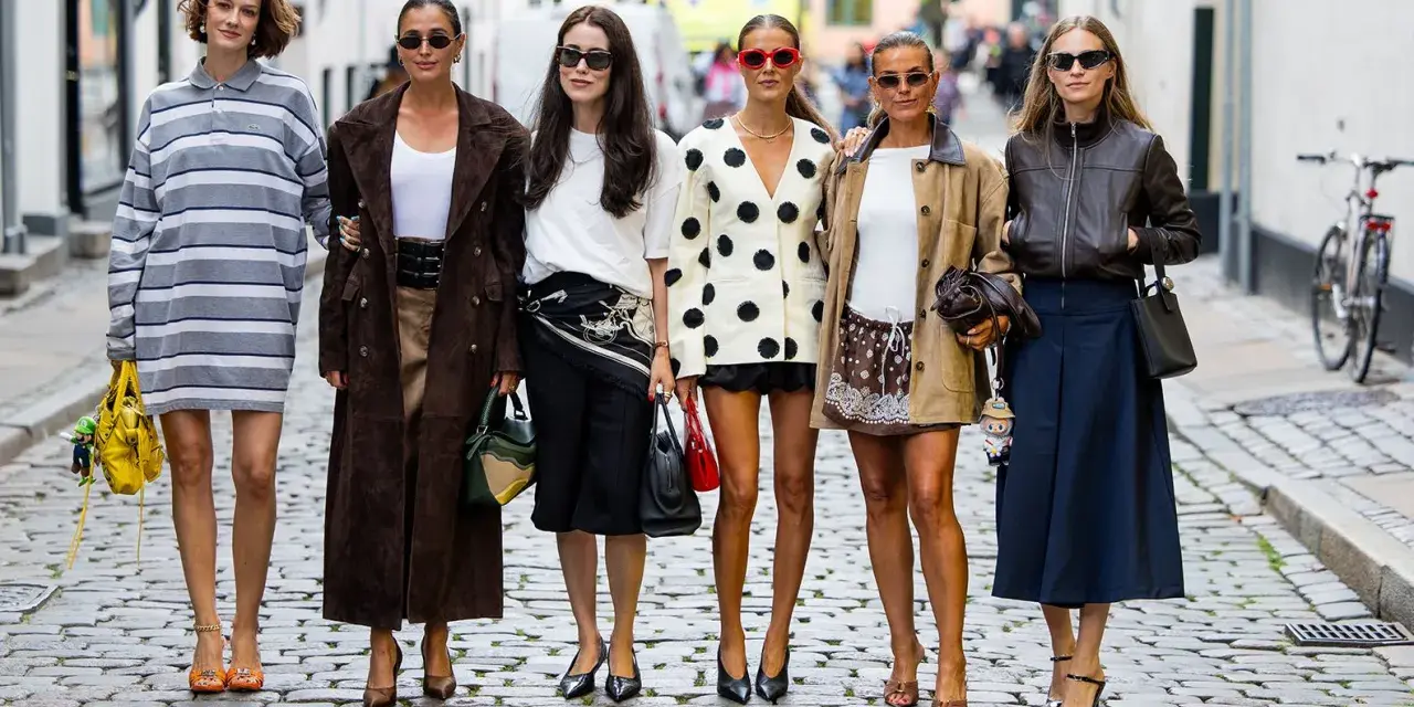 Sechs Frauen in stylischen Outfits, darunter ein gestreiftes Kleid und ein Blazer mit nordischen Mustern, posieren auf einer Kopfsteinpflasterstraße.