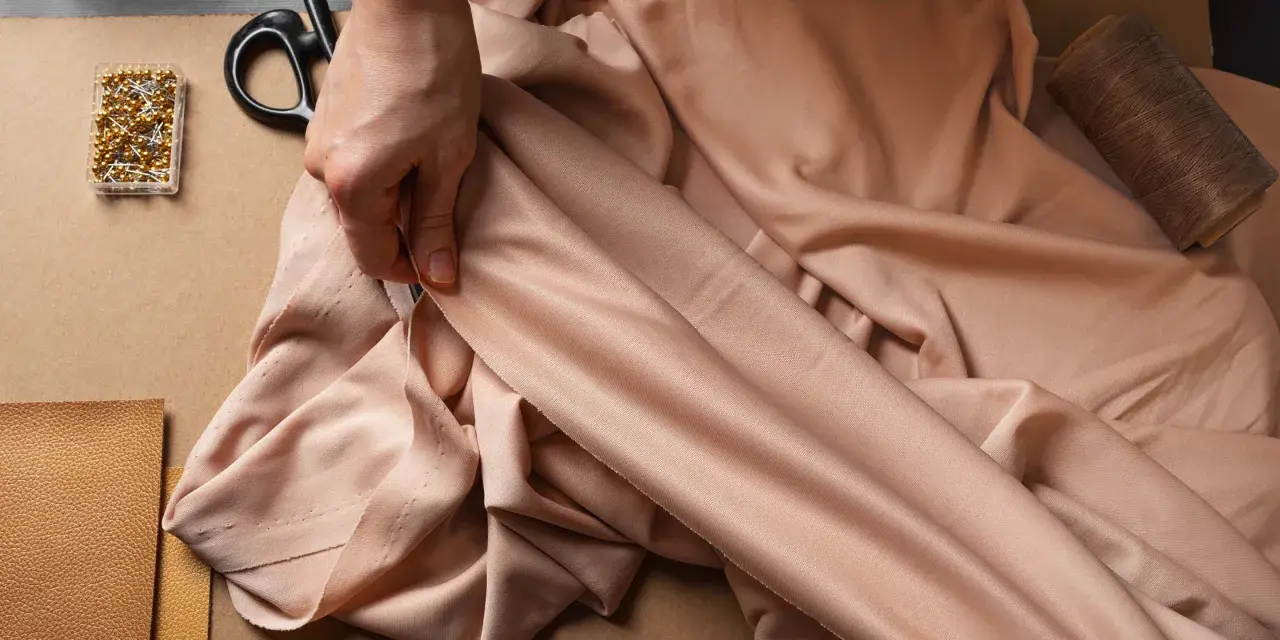 Eine Hand prüft den Fall eines fließenden Stoffes aus Viskose Polyester. Nadeln, Schere und Garn liegen bereit.