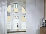 Weiße Schiebegardinen in alten Schienen vor einem Bogenfenster mit Blick auf ein Gebäude. Eine Puppe steht auf der Fensterbank.
