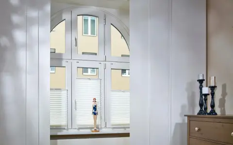 Weiße Schiebegardinen in alten Schienen vor einem Bogenfenster mit Blick auf ein Gebäude. Eine Puppe steht auf der Fensterbank.