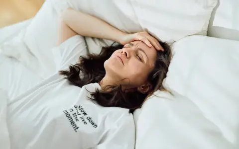 Frau liegt im Bett, Stirn gerunzelt, Hand an der Schläfe. Zu viele Gedanken im Kopf, kein Schlaf möglich.