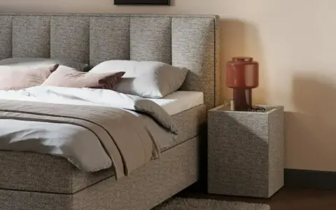 Gemütliches Queensize Bett mit grauem Stoffbezug, Kissen und Decke. Daneben ein Nachttisch mit Lampe.