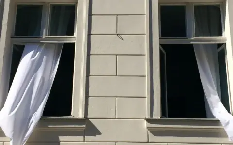 Weiße Gardinen wehen aus zwei offenen Fenstern eines hellen Gebäudes. Die Frage "Gardinen waschen wieviel Grad?" beschäftigt die Hausfrau.