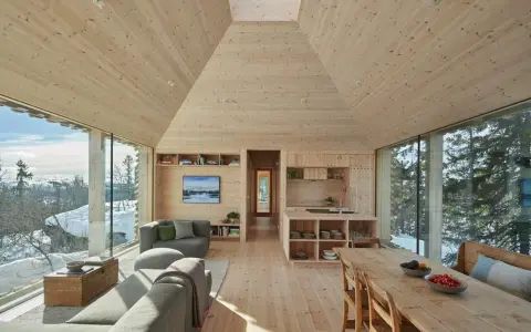 Gemütliches Wohnzimmer mit Holzwänden und Decke, die ein kariertes Muster andeuten. Große Fensterfronten bieten Blick auf verschneite Landschaft.
