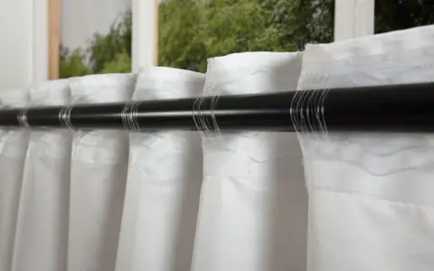 Weiße Polyestergardinen hängen glatt an einer dunklen Stange. Der Stoff fällt in weichen Falten.