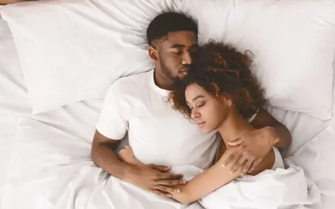 Ein junges Paar schläft eng zusammen in einem Bett. Er hält sie schützend im Arm, während sie sich an ihn kuschelt.