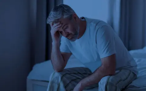 Ein Mann sitzt im Dunkeln auf seinem Bett, die Hand an der Stirn. Er fragt sich, wie lange man ohne Schlaf auskommt.