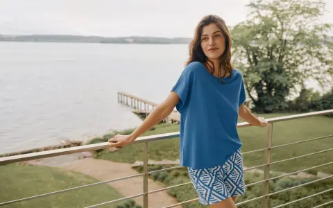Frau auf Balkon mit Blick auf See, trägt blaues Oberteil und gemusterte Shorts. In Modal schwitzt man nicht, es ist angenehm kühl.