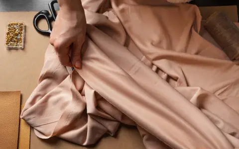 Eine Hand prüft den Fall eines fließenden Stoffes aus Viskose Polyester. Nadeln, Schere und Garn liegen bereit.