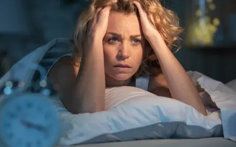 Verzweifelte Frau im Bett, Hände am Kopf, fragt sich, was die Wolfsstunde ist. Ein Wecker ist unscharf im Vordergrund zu sehen.