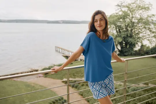 Frau auf Balkon mit Blick auf See, trägt blaues Oberteil und gemusterte Shorts. In Modal schwitzt man nicht, es ist angenehm kühl.