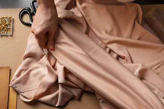 Eine Hand prüft den Fall eines fließenden Stoffes aus Viskose Polyester. Nadeln, Schere und Garn liegen bereit.