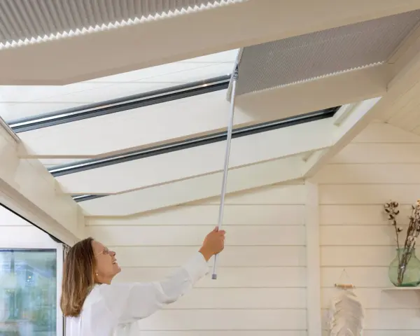 Frau befestigt mit einem Stab eine Faltgardine am Dachfenster im Wintergarten.
