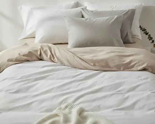 Ein Bett mit weißer und sandfarbener Bettwäsche aus Satin-Baumwolle. Mehrere Kissen liegen auf dem Bett.