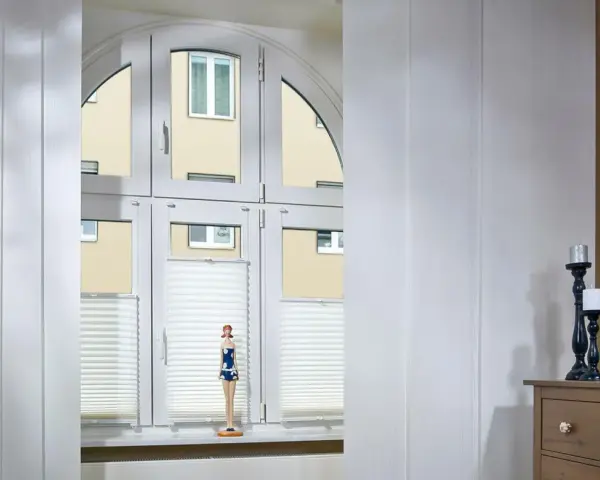 Weiße Schiebegardinen in alten Schienen vor einem Bogenfenster mit Blick auf ein Gebäude. Eine Puppe steht auf der Fensterbank.