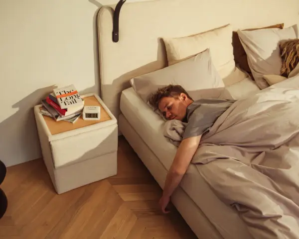 Mann schläft tief und fest auf einem Bett, das sich wie **Kaltschaum** anfühlt. Neben dem Bett liegen Bücher und ein Wecker.