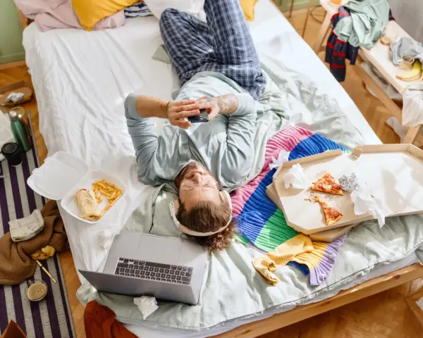 Mann liegt im Chaos auf dem Bett, umgeben von Essensresten und Kleidung. Ein typischer Tag des "bed rotting".