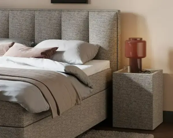 Gemütliches Queensize Bett mit grauem Stoffbezug, Kissen und Decke. Daneben ein Nachttisch mit Lampe.