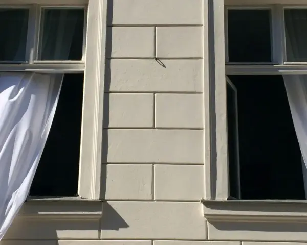 Weiße Gardinen wehen aus zwei offenen Fenstern eines hellen Gebäudes. Die Frage "Gardinen waschen wieviel Grad?" beschäftigt die Hausfrau.