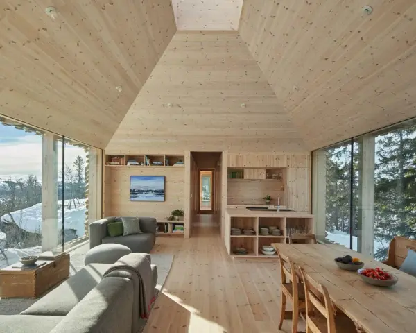 Gemütliches Wohnzimmer mit Holzwänden und Decke, die ein kariertes Muster andeuten. Große Fensterfronten bieten Blick auf verschneite Landschaft.
